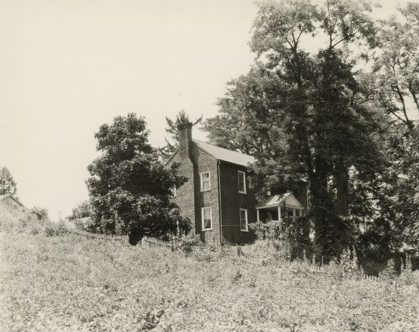 View along side of hill toward the brick manor house with an exterior chimney on the end of the building. A fence is going up the hill in front of the house, and a building is at the top of the hill on the far left.