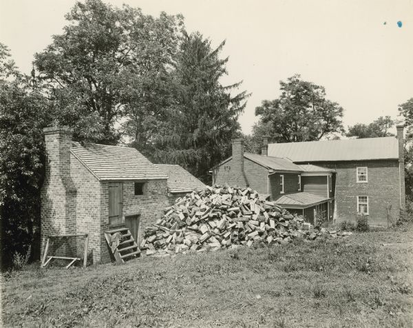 View toward the back of the brick manor house. Another smaller brick building is behind the house on the left. A large pile of fuelwood is against the smaller building.