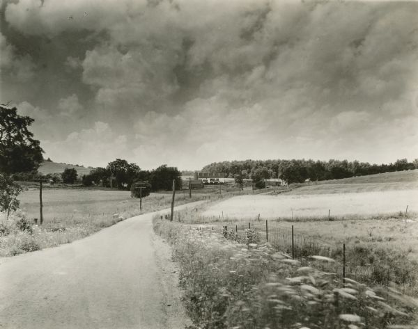 View down road and across fields toward the Walnut Grove farm. The manor house is in the background on the left, a large barn with a silo is in the center. Near the barn on the right is the blacksmith shop and mill building.