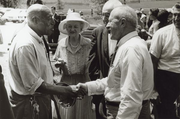 The two men are shown shaking hands with a crowd of people around them. Caption reads: "Grandson Greets Grandson — When Cyrus Hall McCormick perfected the reaper at the family farm in Virginia, Jo Anderson toiled with him in the blacksmith shop to build it. One hundred-thirty-five years ago, at the first successful test on the reaper, Jo Anderson walked beside the machine, raking the platform clear of the cut grain. Last month a portion of the Walnut Grove Farm was designated a national landmark by the National Park Service. One of the highlights of the day was the reunion of the grandsons of those two men — former IH Board Chairman Fowler McCormick, grandson of Cyrus Hall McCormick, and Harry Wilson, grandson of Jo Anderson. Mr. Wilson will retire soon, after almost 50 years' service as custodian of the site."