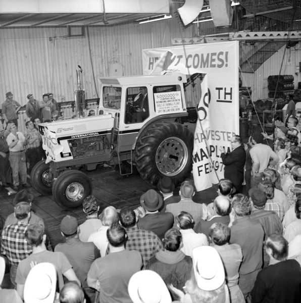 The 5,000,000th International Harvester tractor to roll off the assembly line on Feb. 1, 1974. The tractor is a Farmall 1066. Slightly elevated view of a man driving the tractor through a large banner surrounded by a crowd of people.