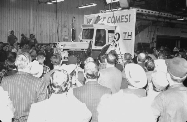The 5,000,000th International Harvester tractor to roll off the assembly line on Feb. 1, 1974. The tractor is a Farmall 1066. Side view over the heads of a crowd toward a man driving the tractor through a large banner.