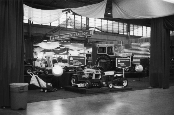 View toward a display set up in a factory. A sign hanging above the display reads: "Five Millionth Tractor." A sign behind a Cub Cadet reads: "IH International Harvester, Presenting the 500,000th Cub Cadet. ...a NEW RECORD for lawn and garden tractors."