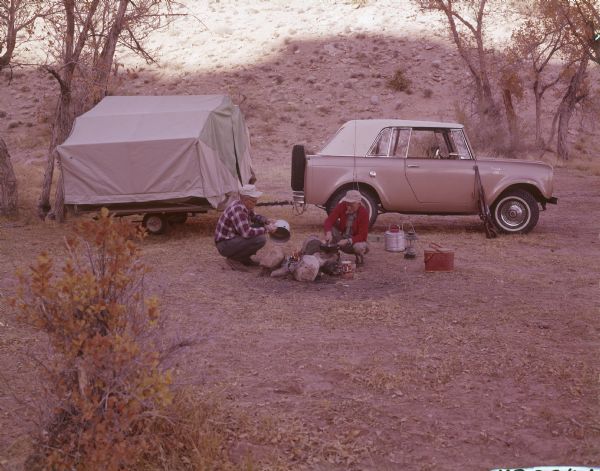 Two men are cooking at a campfire. Behind them is a parked Scout truck which has a trailer attached to the back. Two rifles are leaning against the side of the Scout just in front of the passenger door.