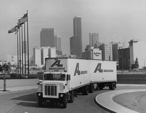 The International 4100 Conco on a curved road with tall buildings in the background. News Photo text reads: "New International Transtar 4100 Conco model combines the best features of both conventional cab and cab-over trucks. The 76-inch wide aluminum cab is positioned to the left which provides driver room similar to that of a 96-inch wide cab-over model. The new Conco has been designed primarily as a fleet tractor which can be used for pulling relay doubles, a 45-ft. trailer or 'day and night service' combination line haul and pickup and delivery."