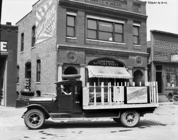 View across street towards a man sitting in the driver's seat of a delivery truck for the Hur-Mon brand of soda. A spare tire is mounted on the roof of the cab of the truck. The storefront behind the truck has a sign above the window that reads: "Cedar Rapids Bottling Works" and in the alley on the left is a small loading dock for the store. A storefront next door on the right sells tires and gas.