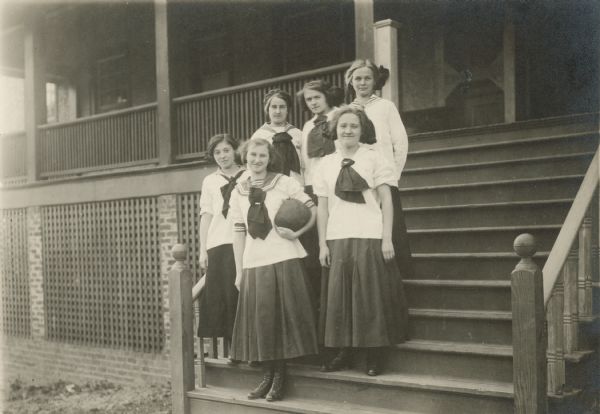 Group portrait of five girls posing on the steps of a building. They are wearing uniforms of white blouses with striped collars and dark neckties over dark skirts. The girl in front is holding a basketball in her arm.