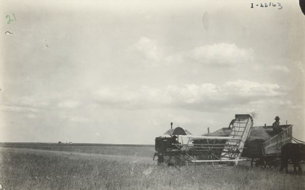 Man using motor cultivator in a field. Two men are standing in a horse-drawn wagon nearby. In the distance across the field are two buildings.