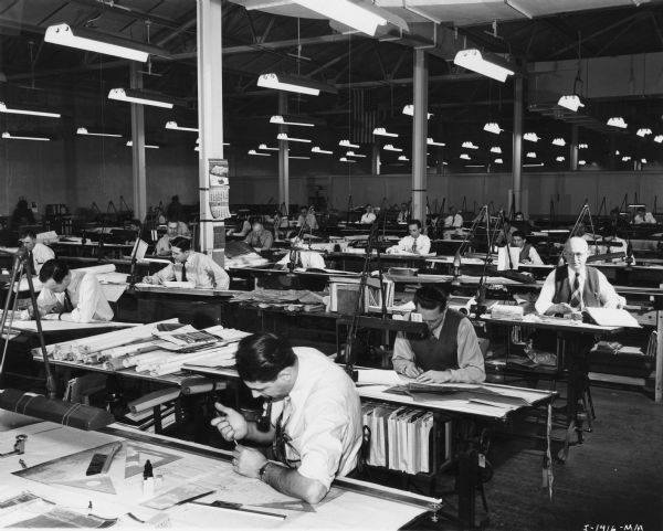 A group of men working at tables in the Drafting Department at International Harvester's Tractor Works. An American flag is hanging from the high ceiling in the back of the room.