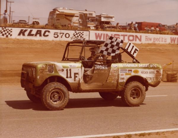 Driver's side view of an International Scout SS-II on a racetrack. In the background are spectators watching the race from behind a fence.