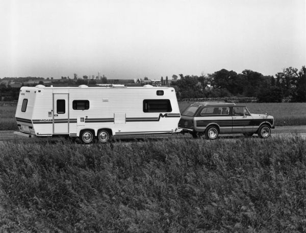 View toward an International Scout Traveler pulling a trailer. News Photo sheet reads: "The 1979 International Scout Traveler can tow trailers weighing up to 5,000 lbs. The Traveler's long 118-in. wheelbase adds stability at highway speeds. A new optional auxiliary oil cooler for the automatic transmission is particularly useful for trailer-towing applications."