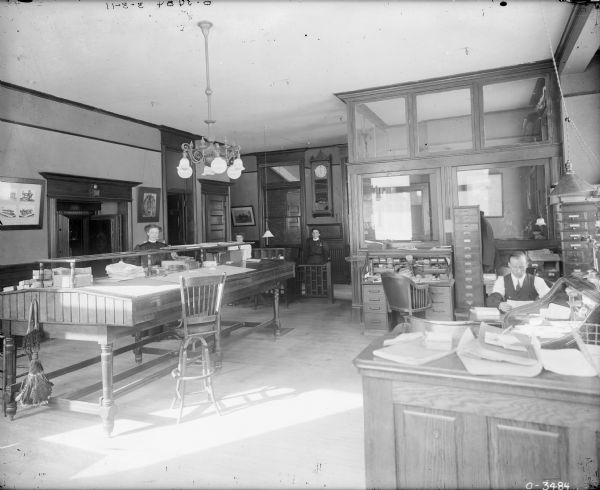 View of four people in a large, wood-panelled office with a high ceiling. One woman is sitting on the left behind a large desk in front of a safe built into the wall. Another woman is sitting behind her. Another woman is standing behind a gate in the center near a Watchmans Electric Clock on the back wall. In the foreground on the right is a roll-top desk with a telephone on top, and sitting at a desk behind is  a man wearing sleeve-garters.