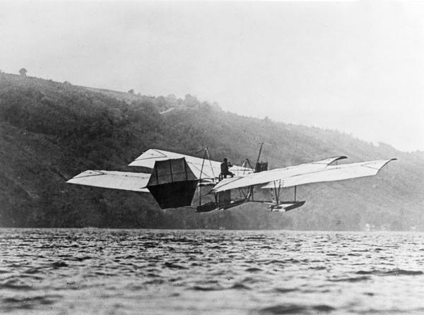 Image of Curtiss flying an airplane over a body of water. There is a hill along the shoreline in the background.