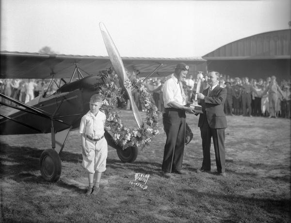 Pilot Orland G. Corben accepts the winner's trophy at the Kroger Air Show in Madison from Kroger Manager D.V. McPeek.  Also in the photograph is Tommy Haines. Corben, manager of the Madison Airport and manufacturer of Corben planes and kits, finished first in the fifteen-mile race, flying one of his own Baby Ace airplanes. A Baby Ace was also featured in the stunt flying at the air show by local aviator Merle Buck.