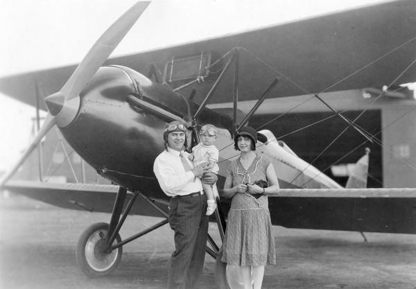 Family portrait, probably taken at Alexander Field in Wausau. Included are an unidentified aviator, his wife, his baby, and a Waco 10 aircraft. Goggles, which were necessary to protect pilots from the wind in open cockpits, are emblematic of early aviation.