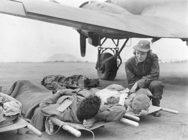 At Iwo Jima Airfield #1 Gwen Jensen, a flight nurse, talks with wounded Marines who are about to be evacuated by airplane. Mount Surabachi, from which the U.S. flag was first flown ten days before, is in the background. This photograph is one of many taken by Milwaukee photographer Dickey Chapelle during the prolonged battle for Iwo Jima. She was present within nine days of the first Marine landing.