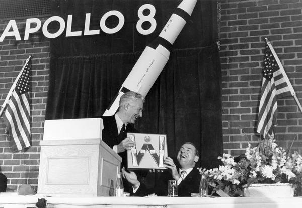 Astronaut James A. Lovell accepting a gift of cheese from Wisconsin Governor Warren P. Knowles during a gathering in Lovell's honor. A model of the Apollo 8 in which Lovell had recently orbited the Moon with Frank Borman and William Anders decorates the podium.  
