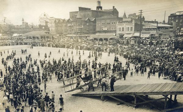 Elevated view of crowd gathered on the beach at Atlantic City to watch a Curtiss airplane being pushed on an inclined ramp leading to the boardwalk.