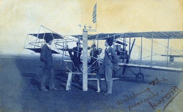The Curtiss Pusher owned by Arthur Pratt Warner of Beloit, the first man to fly in Wisconsin and the first person to purchase an airplane.  Slightly below the flag is the "Aero-meter," Warner's invention for measuring wind speed. The April 3, 1910 postmark on the reverse side of the photographic postcard proves that Warner exhibited his plane at the Los Angeles Air Meet.