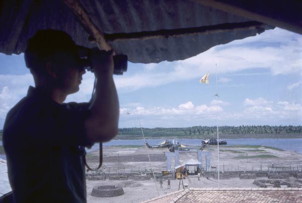 An unidentified image from the collection of Dickey Chapelle, a freelance photographer, shows a U.S. Marine on duty in the watchtower of a South Vietnamese base. Behind him are several Piesecki CH21-C helicopters and the perimeter gate. In the distance are a river and the Vietnamese landscape.