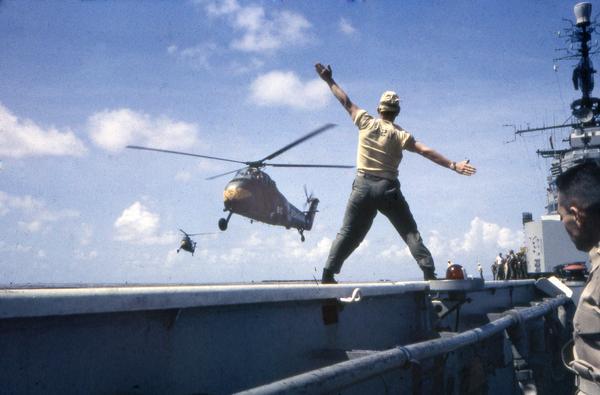A crewman on a U.S. Navy aircraft carrier signals to the pilot of an incoming U.S. Marine Corps CH-34 Choctaw helicopter. The landing was photographed by Milwaukee photographer Dickey Chapelle during the 1950s.