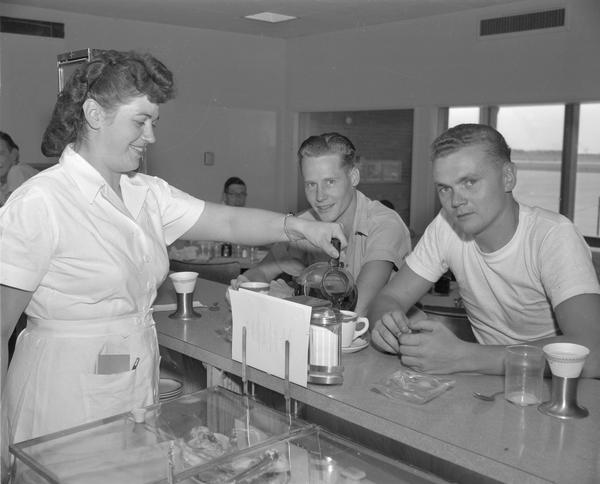 The lunch counter at Truax Field, formerly Madison Municipal Airport.  Two men are sitting at the counter, probably airport employees, and a waitress is pouring coffee.
