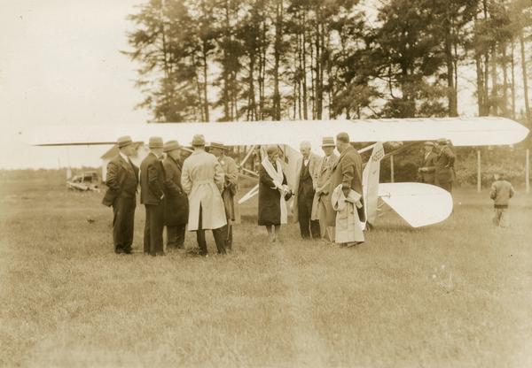 Walter J. Kohler, Sr., (partially hidden, wearing a straw hat and with a flower in his lapel) makes a stop to which he had flown in his airplane, the "Village of Kohler."  Kohler's plane was a Mahoney-Ryan-Brougham, a commercially available aircraft patterned after the "Spirit of St. Louis." Kohler was Wisconsin's first aviation-minded governor.  He campaigned for office using the airplane in 1928 and during his single term in office he frequently flew between the capital and his home in Kohler. Although unidentified, this photograph is believed to have been taken during the 1928 campaign.  The woman in the group is Charlotte Kohler.