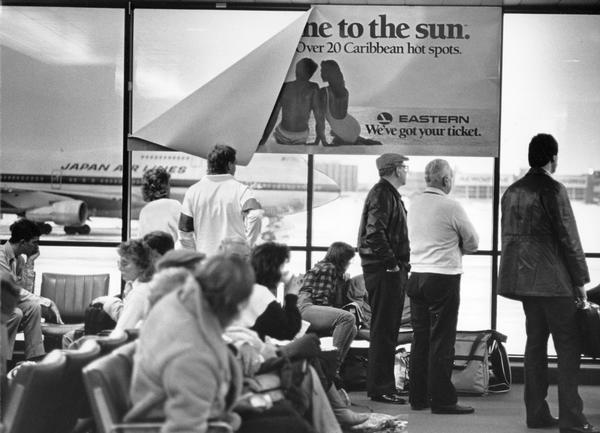 Bored travelers at General Mitchell International Airport, apparently taking their cue from a droopy travel poster advertising vacations in the sunny Caribbean. The Japan Airlines 747 in the background had been diverted from O'Hare after a snowstorm closed the Chicago airport. The 747 series, which was introduced by Boeing in 1970, is one of the most successful planes in aviation history. In 1999 the 747 received its own U.S. postage stamp, recognizing its place as one of the top three aviation achievements of the century-along with the Wright Brothers and Lindbergh's trans-Atlantic crossing.
