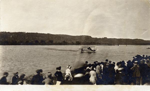 A crowd gathered on the Mississippi River shoreline near Prairie du Chien. They are watching pioneer aviator Hugh Robinson taxi his Curtiss flying boat.