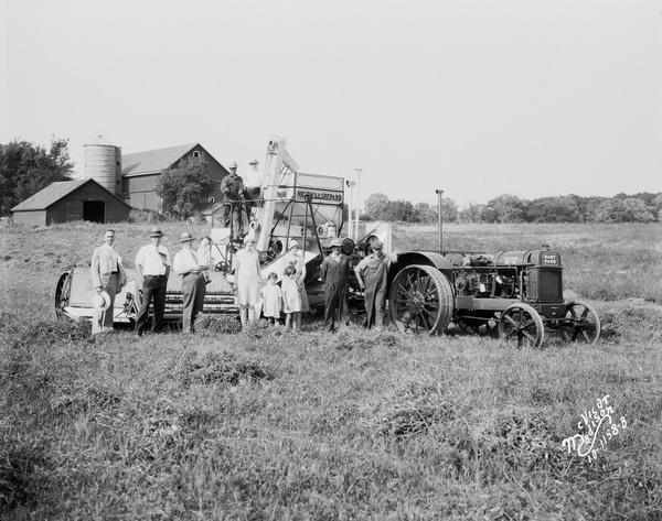 A group portrait of men, women and children standing in a field, with a Nichols & Shepard combine thresher and Hart Parr tractor behind them. In the background are farm buildings, including a silo and barn.