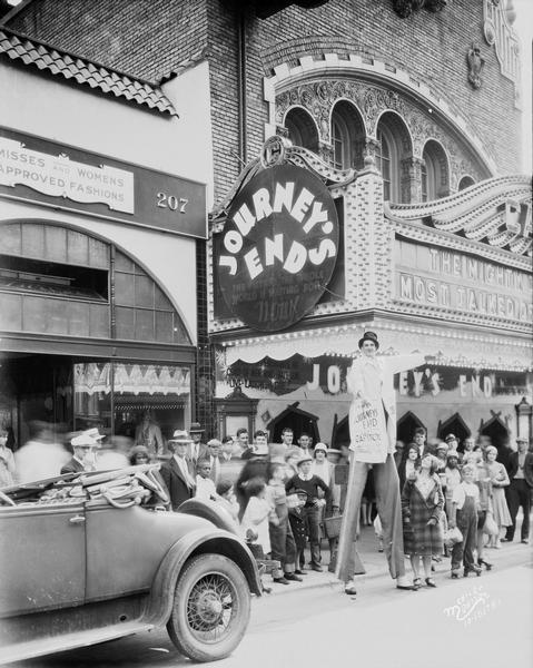 View from street of the facade of the Capitol Theatre at 209 State Street. The marquee is advertising the movie, "Journey's End." There is a crowd on the sidewalk in front of the theatre, and a man on stilts is standing in the street near an automobile.
