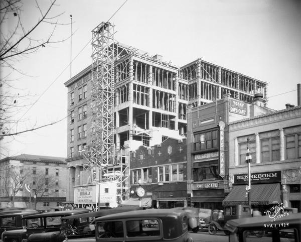 Construction of an addition to the State Bank of Wisconsin at 1-5 West main Street, as viewed from across West Main Street, with automobiles angle parked in foreground. In the background are buildings, including Woolworths at 1 East Main Street, Badger Candy Kitchen at 7 West Main Street, E.W. Parker Jewelers at 9 West Main Street, Cop's Cafe at 11 West Main Street, and Rennebohm Drug Store #3 at 13 West Main Street, which was part of the Levitan Building.