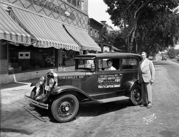 Chevrolet automobile advertising the Chevrolet Six Economy Drivers' contest with "Test your driving skills, economy drivers' contest, $75 in cash prizes, free, ask driver for information, Hult's Capital Garage" painted on the driver's door. Cedric Parker is sitting at the wheel, and Ralph A. Hult is standing beside the car. The car is parked in front of Hult's Garage which was located at 608-612 East Washington Avenue.