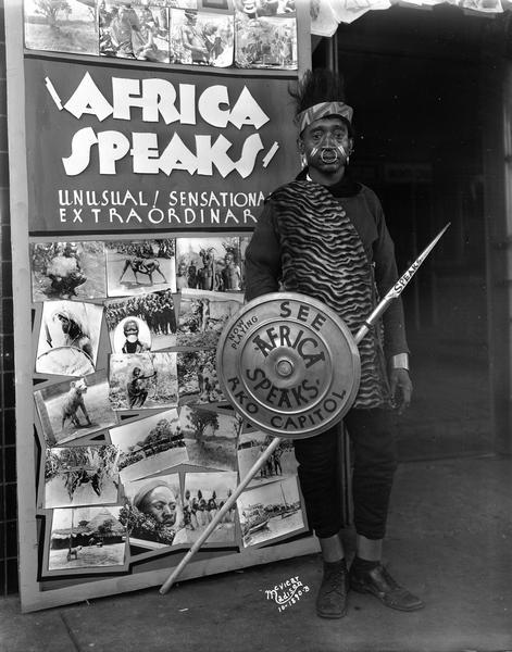 Man dressed as an African standing beside poster advertising the movie, "Africa Speaks," in front of the Capitol Theatre, 209 State Street.
