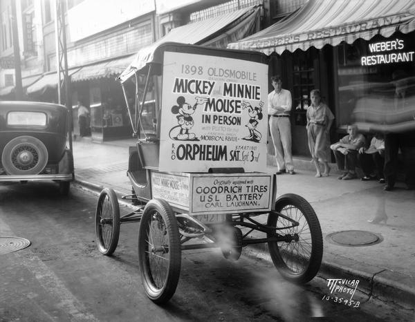 1898 Oldsmobile decorated with advertising for "Mickey and Minnie Mouse in Person" at RKO Orpheum Theatre in front of Weber's Restaurant, 218 State Street. Included are advertisements for Goodrich tires, USL Barpy, and Carl Laughan.