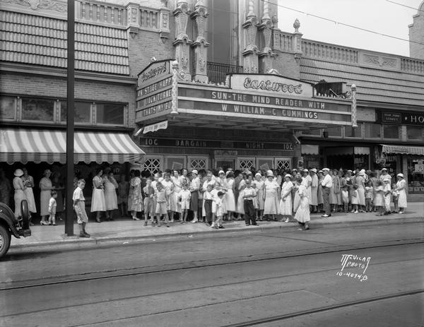 View from street of the Eastwood theatre, located at 2090 Atwood Avenue with  "Sun - The Mind Reader with W. Williams and C. Cummings" and "Happy Jack Turner - That's My Boy" on the marquee. A crowd of people is waiting for the doors to open. The other storefronts on the block are the Harold F. Huss sandwich & soda shop, Obert Brickson Jewelry store, and the Hommel Brothers Star Food grocery.