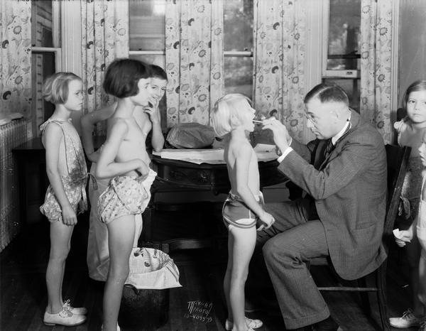 Doctor John E. Gonce examining a child's throat while other children wait in line, in order to go to "The Capital Times" Kiddie Camp.