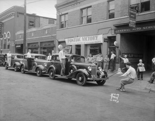 Arthur Santell is pulling three Pontiac automobiles, each with a man on the running board, with his teeth in front of General Auto Service and Henry Motor Co., 323 West Johnson Street.