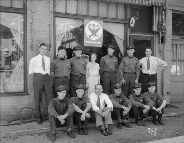 Group portrait of 13 Goodyear Service employees. Includes one woman, and nine men in uniform, posing in front of a store window with an NRA (National Recovery Administration) sign, 205 N. Bassett Street.