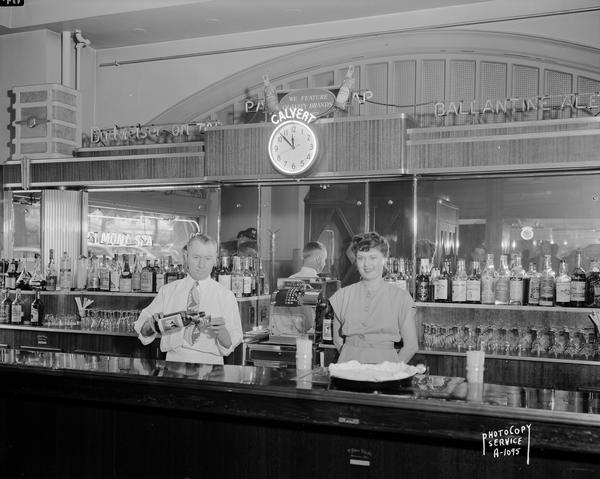Richard and Ione Edwards behind the bar at the Belmont Spa, in the Belmont Hotel, 31 North Pinckney Street, with a clock advertising Calvert liquor.