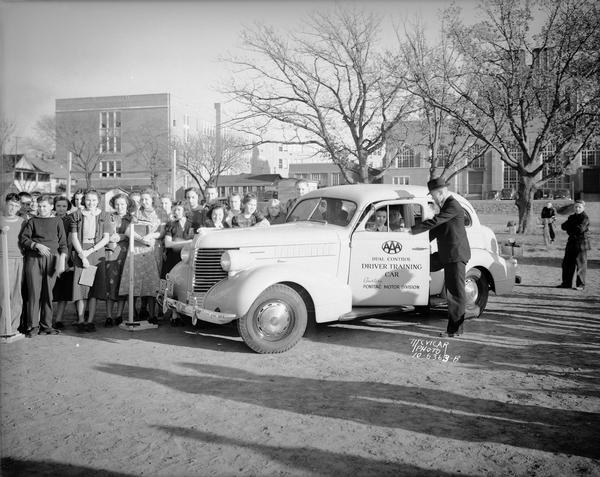 AAA dual-control training car parked at East High School, 2222 East Washington Avenue. Several students are in the car, a Pontiac, with a man standing next to it and a group of students, while a group of students look on.