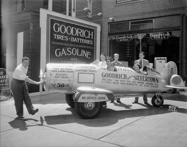 A man spinning the propeller with Frank A. Sloan in the pilot seat and a women is sitting in the back of a car that looks like an airplane. They are at a Goodrich Silvertown Stores, 515 University Avenue to advertise tires. The car/airplane has the following sentence painted on the side: "The only tire with the life-saver golden ply, three times safer from blowouts at high speeds."