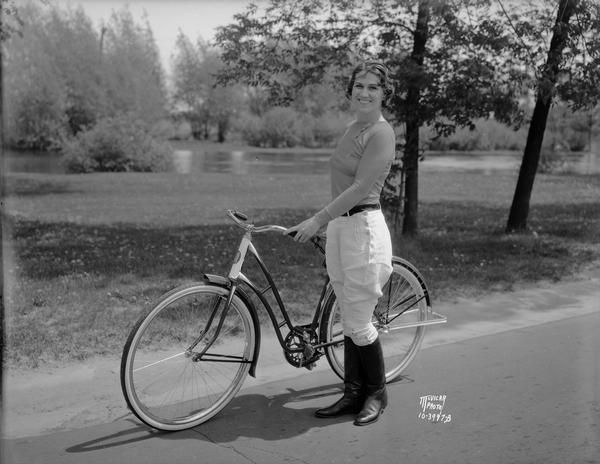 Janette Serrec, lecturer and writer on the "Fine Art of Successful Living," standing beside a Hawthorne bicycle from Ward's. She is wearing jodhpurs and boots.