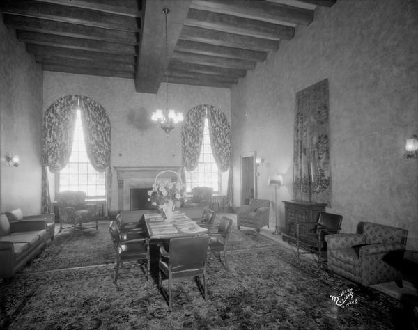 First Congregational Church student lounge, at 1609 University Avenue. The room is furnished with chairs and couches along the walls, two large carpets, and a table with chairs in the center of the room. Along the back wall are two tall windows with curtains flanking a fireplace. The wood ceiling has a chandelier hanging above the fireplace.