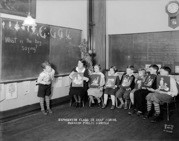 A teacher and seven students are holding pictures in "Expression class" in a deaf classroom at Doty school, 351 W. Wilson Street. A boy is standing in front of a chalkboard in front of the class. Written on the billboard is: "What is the boy saying?"