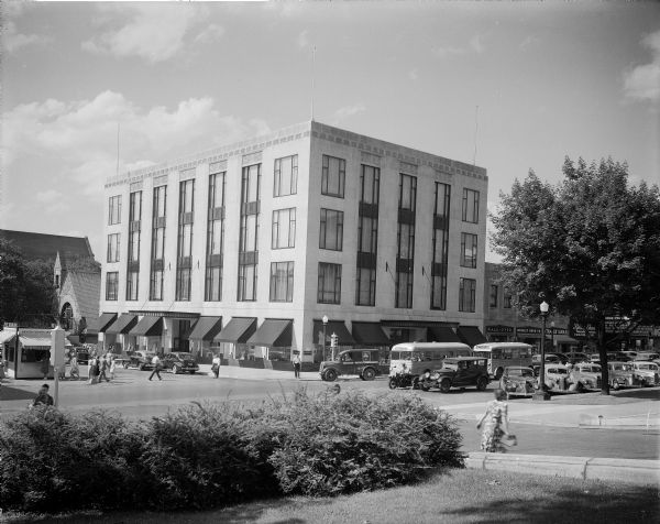 Harry S. Manchester's, Incorporated, department store on Capitol Square, 2-6 East Mifflin Street. View of people, buses and automobiles at the intersection of East Mifflin Street and Wisconsin Avenue. Also in view are the Madison Business Association information booth, Unitarian Society meeting house on Wisconsin Avenue, Walk-Over Shoe Store, Moseley Book Co., Strauss Shoes, Kinney Shoes, and the Strand Theater on East Mifflin Street.