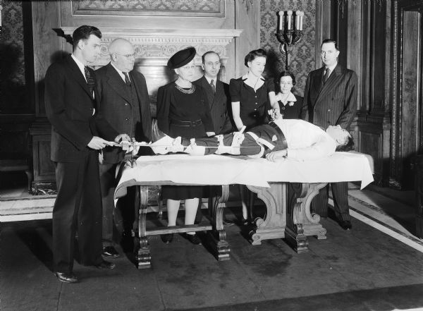 State government employees are being given a demonstration in splinting technique by Red Cross instructors in the Wisconsin State Capitol Assembly parlor. The victim on the table, E.C. Giessel of the Budget Bureau, is surrounded by, l to r: Miles F. Fenske and C.N. Maurer, instructors, of the Motor Vehicle Dept.; Ica Faber, Unemployment Compensation; G.A. Lofye, Secretary of State's office; Virginia Siebecker, Dept. of Public Instruction; Elsie M. Wood, Revisor of Statutes Office; and Clyde S. Tutton, instructor from the Beverage Tax department. Photograph published in "Wisconsin State Employee Magazine," April 1942.