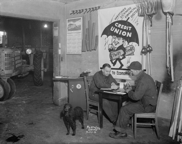 Claire Onsgard, treasurer of the Dane County Farmers Equity Union Coop Credit Union in Cottage Grove showing member Fritz Swenson where to sign his loan application.  They are sitting at a table in front of CUNA poster with tractor in background and dog in foreground.