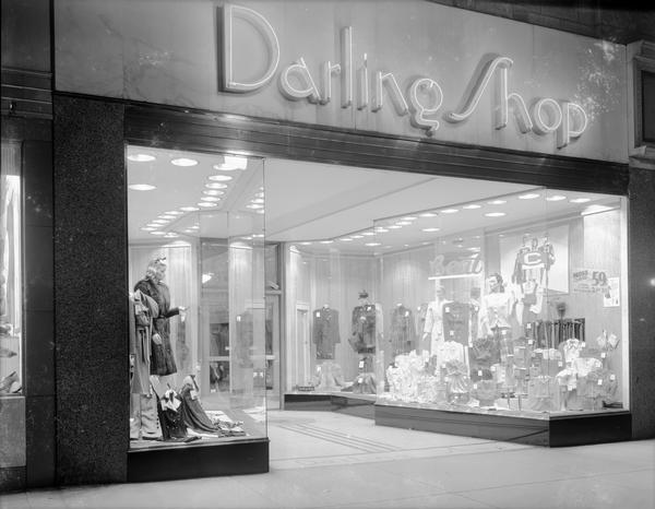 Exterior night view of Darling Shop, 9 East Main Street, with display windows and lighted neon "Darling Shop" sign.