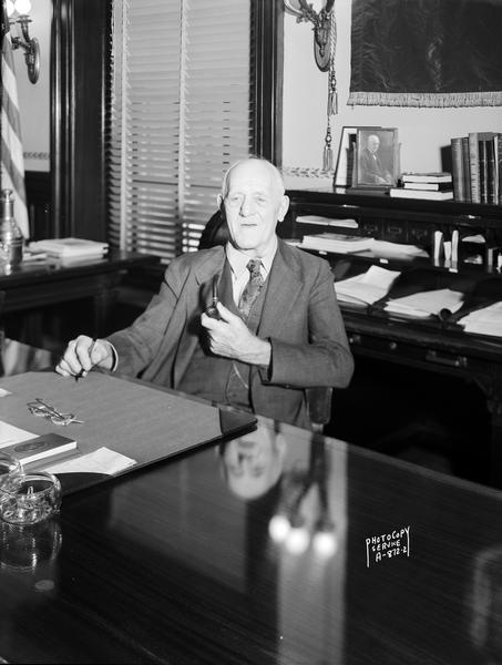Wisconsin Governor Walter S. Goodland sitting at his desk in the Capitol with pipe in hand.